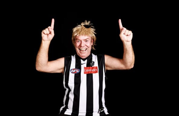 man smiling in blonde mullet wig in black and white striped guernsey with arms raised in victory on black background