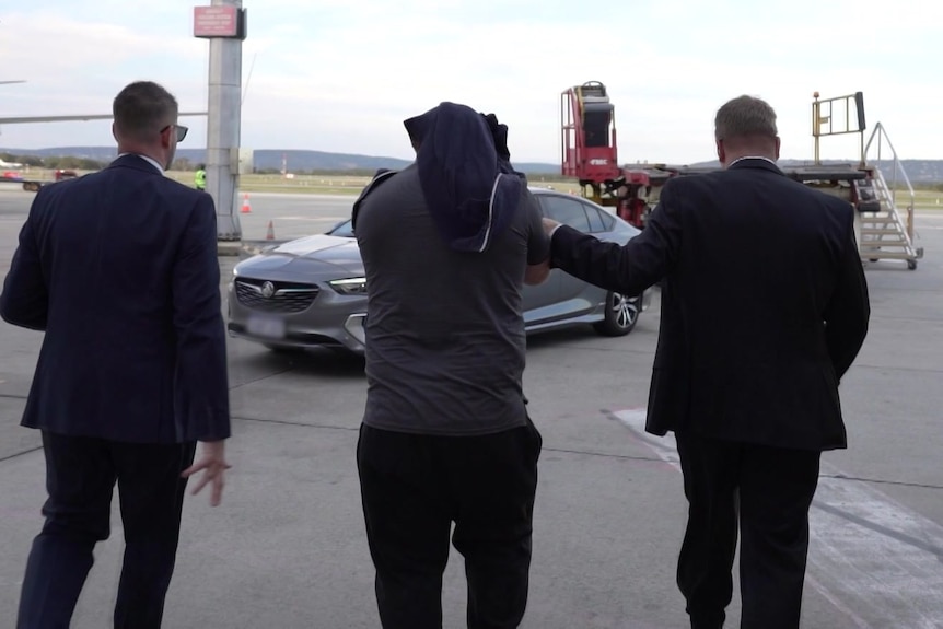 Three men pictured from behind walk on a tarmac towards a car