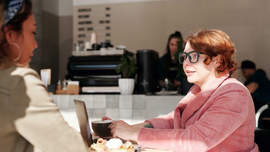 Two women chat at a cafe in a story about when is the right time to talk about pay salary when going for a job.