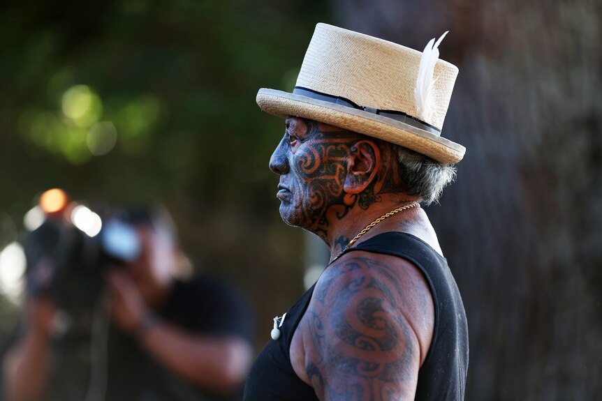 A Māori man with Tā moko face tattoos, seen in side profile, wearing a black singlet and hat