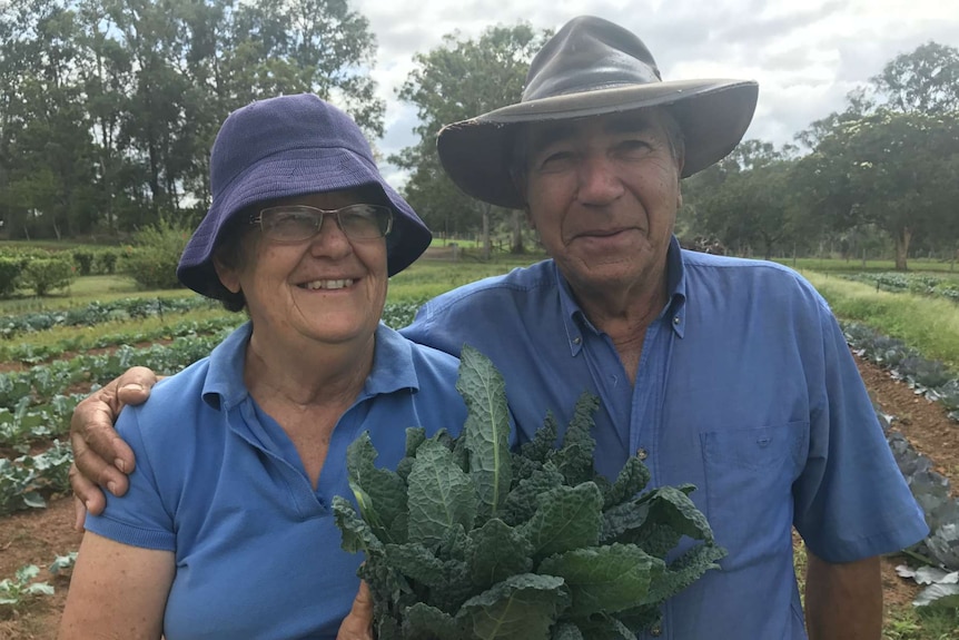 Bruno has his arm over Trish's shoulder as she holds up freshly harvested Asian greens.  They are smiling at the camera.