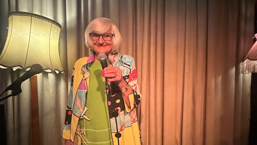 A lady with a blonde bob wearing glasses and holding a microphone looks at the camera smiling.