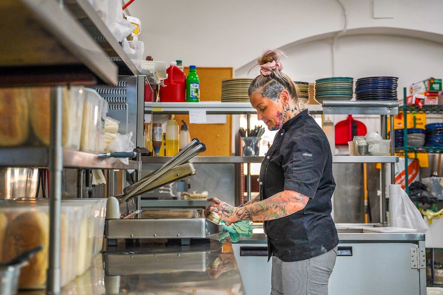 A woman with tattoos, a black shirt, and grey jeans, works in a kitchen.