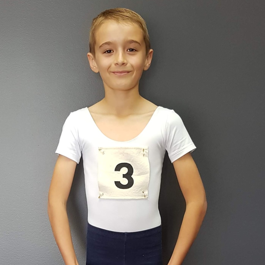 A boy with short, fair hair stands smiling, wearing a white t-shirt, black pants, and the number three pinned to his chest.