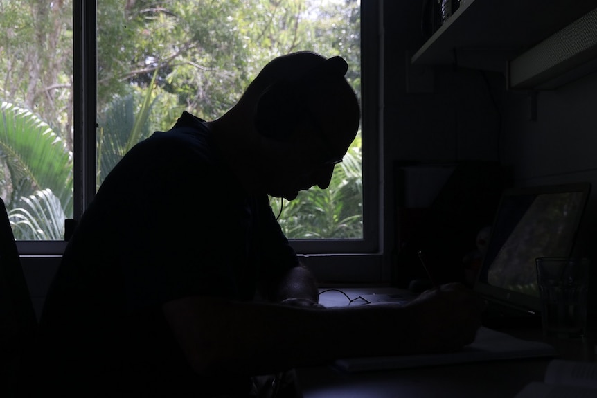 Silhouette of middle-aged man sitting at desk and typing on computer against bright window backdrop.