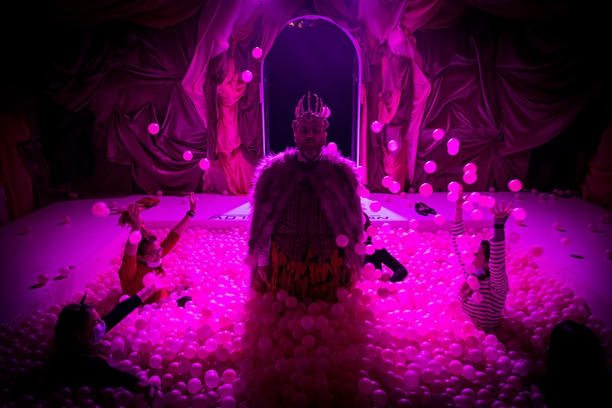 Photo of actor Drew Fairley standing in a pink light lit ballpit with audience members during a A Midnight Visit scene.