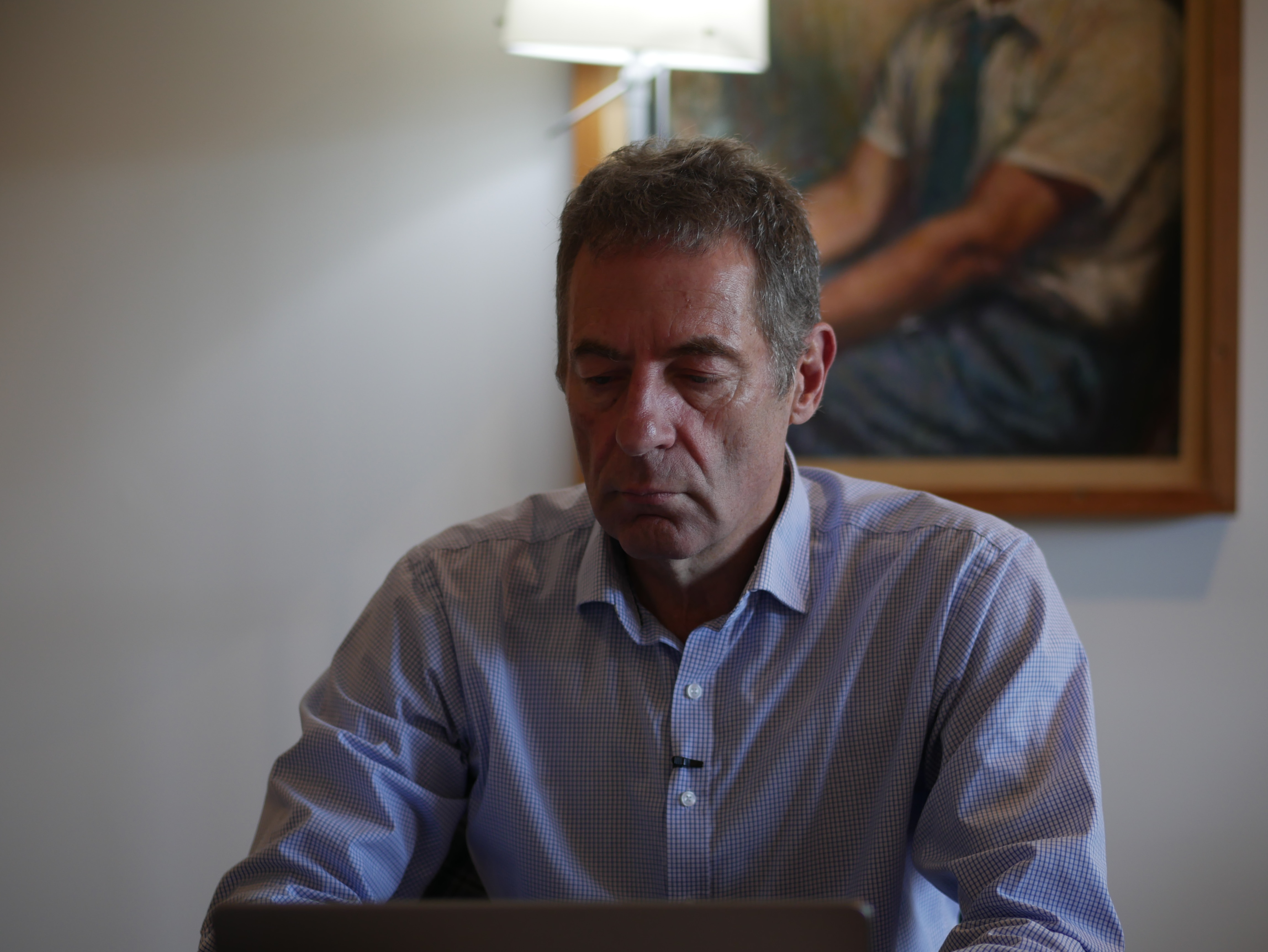 Man seated at table looking down at something, with a light in the background casting a halo