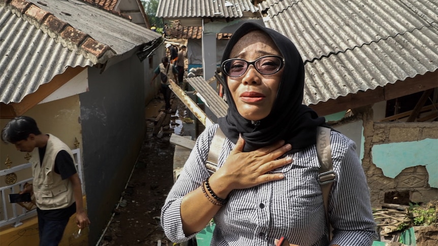 A woman wearing a striped top and dark blue head covering with sunglasses stands outside a damaged house