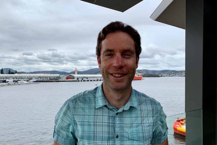A man in a checkered blue shirt looking at the camera and smiling, Hobart's waterfront is behind him