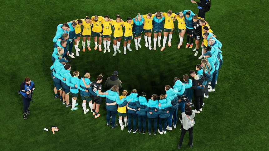 A group of Matildas players and coaches form a heart on the pitch as they talk after a big match.
