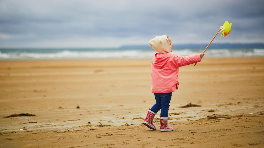 A little girl in gumboots and winter jacket is playing on a beach with a wind wheel.