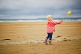 A little girl in gumboots and winter jacket is playing on a beach with a wind wheel.