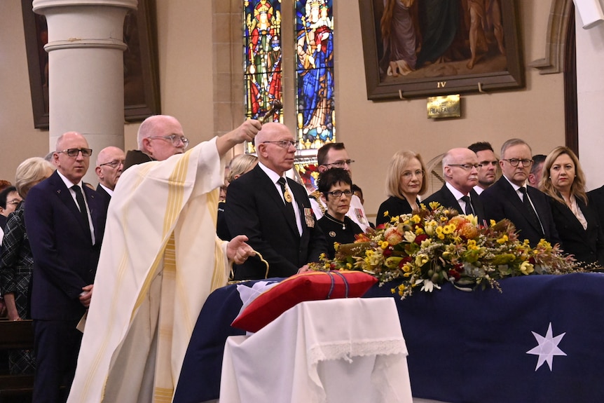 a coffin draped in an australian flag, beside a priest and row of dignitaries 