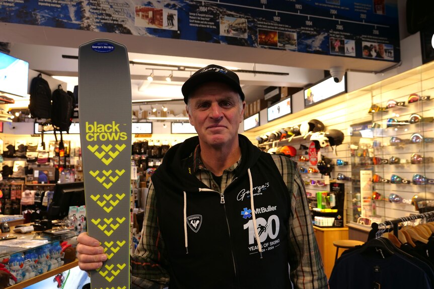 A man wearing a black hoodie that says 100 years of skiing on it, he is holding a pair of skis.