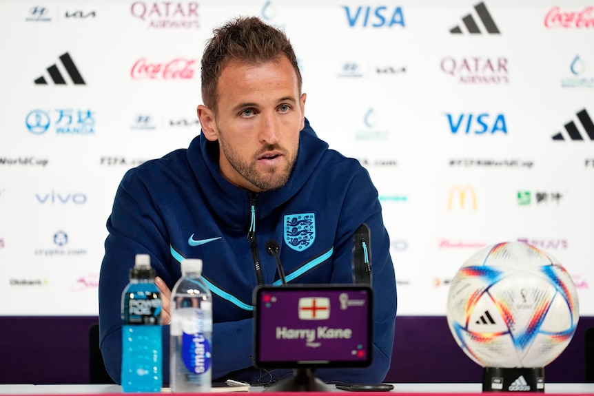 Harry Kane sits at a table behind a wall of world cup logos. 