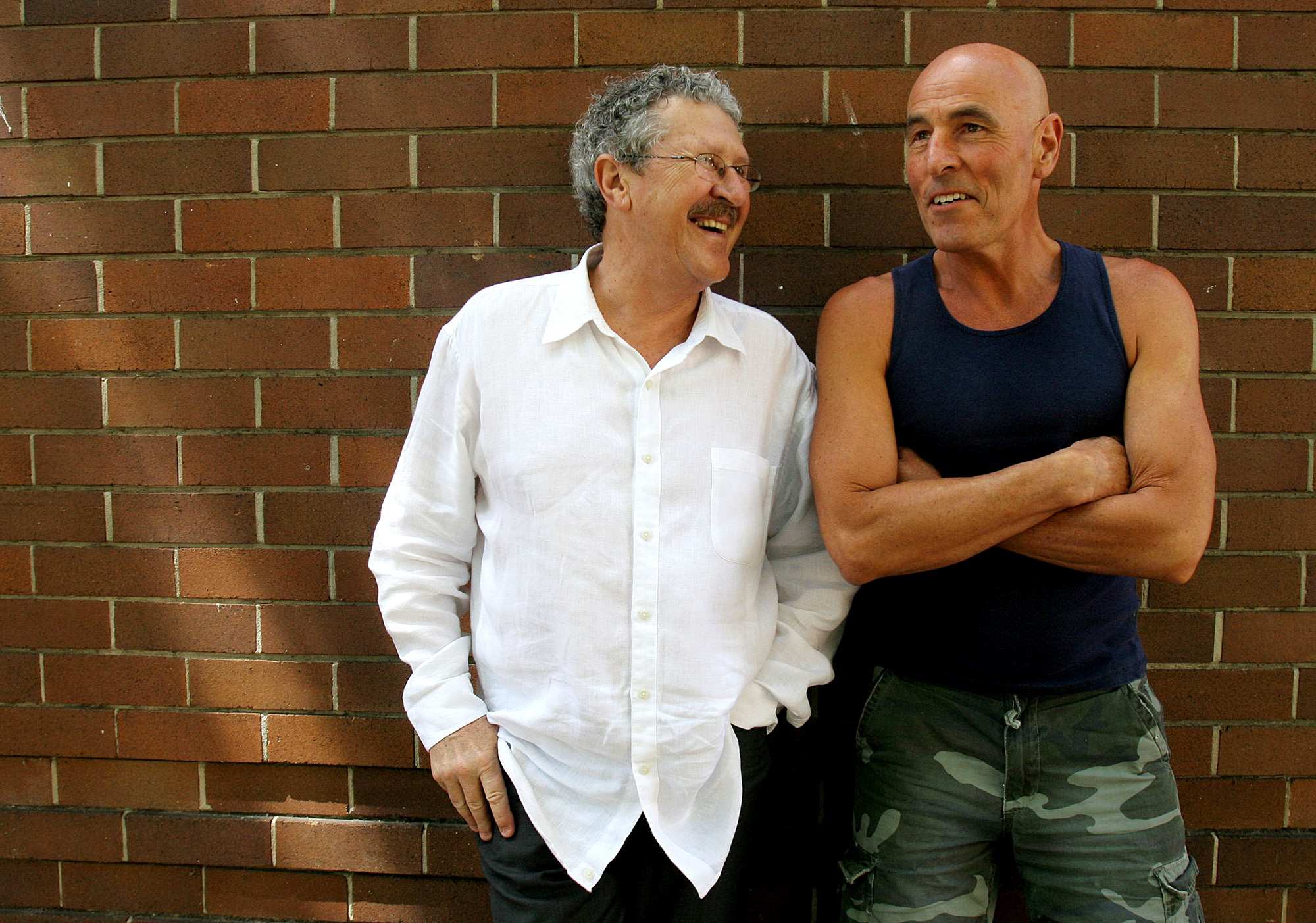 A man with glasses and grey hair laughs with another man who stands with his arms crossed leaning on a brick wall