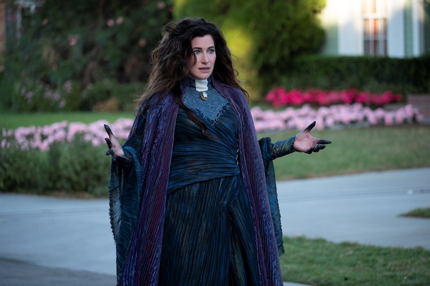 A white woman in her late 40s with long brunette hair wearing a purple witch outfit stands with hands out darkened with ink