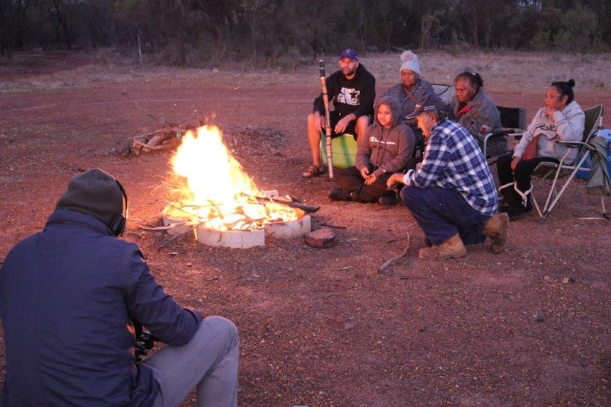 A man squats down as he films six Aboriginal people around a campfire as the sun sets.