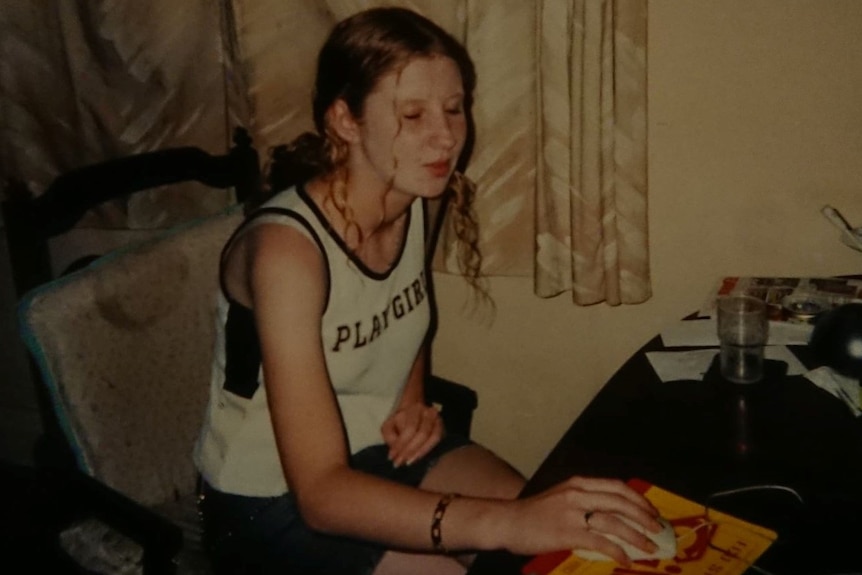 A teenaged girl wearing a top that says "playgirl" sits at a desk with her hand on a computer mouse.