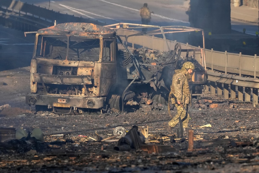 A Ukrainian soldier walks past debris of a burning military truck, which is scattered across an ashphalt street.