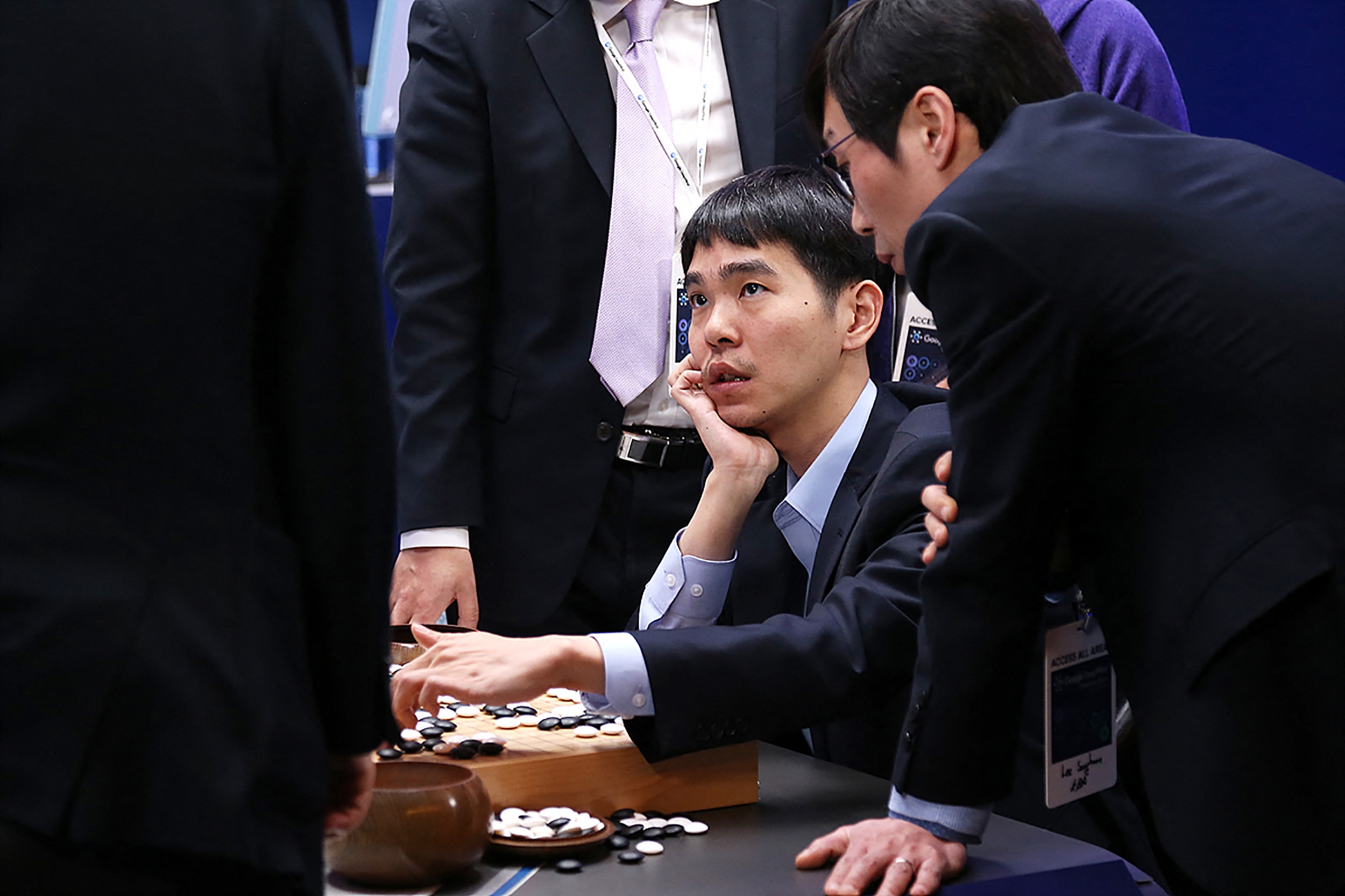 A man in a blue jacket and collared shirt sits at a table with a Go board and pieces of black and ivory white.