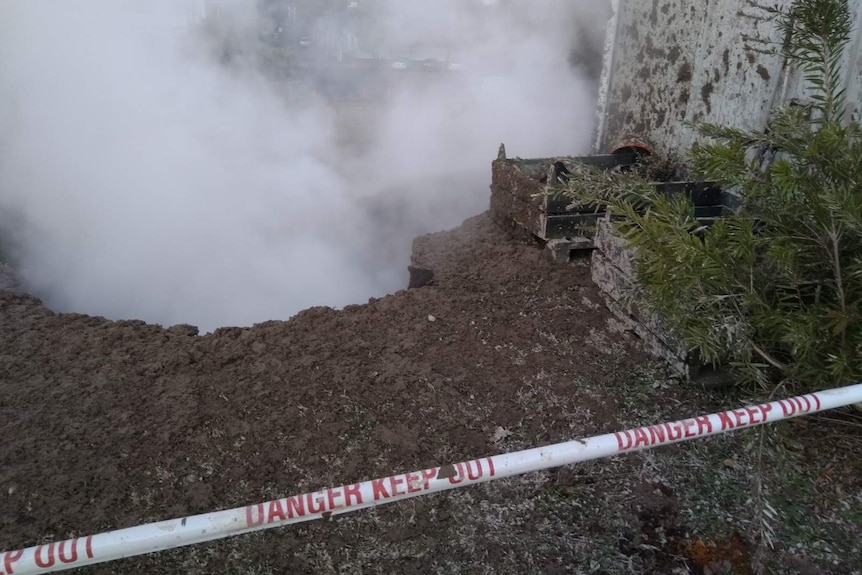 Steam rises from a pit, which is coated with splats of mud around the edges. There is safety tape around the hole.