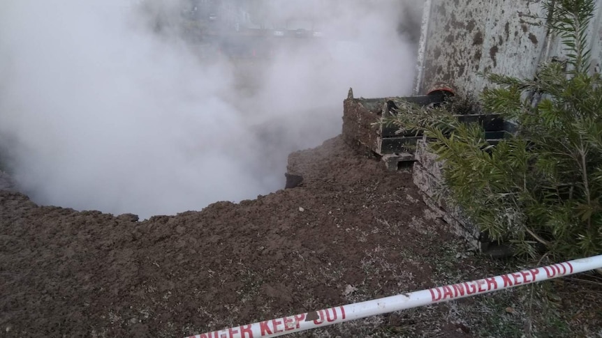 Steam rises from a pit, which is coated with splats of mud around the edges. There is safety tape around the hole.