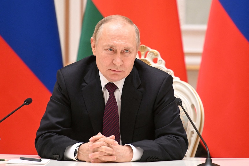 Vladimir Putin sits at a desk with microphones in front of him and flags in the background. 