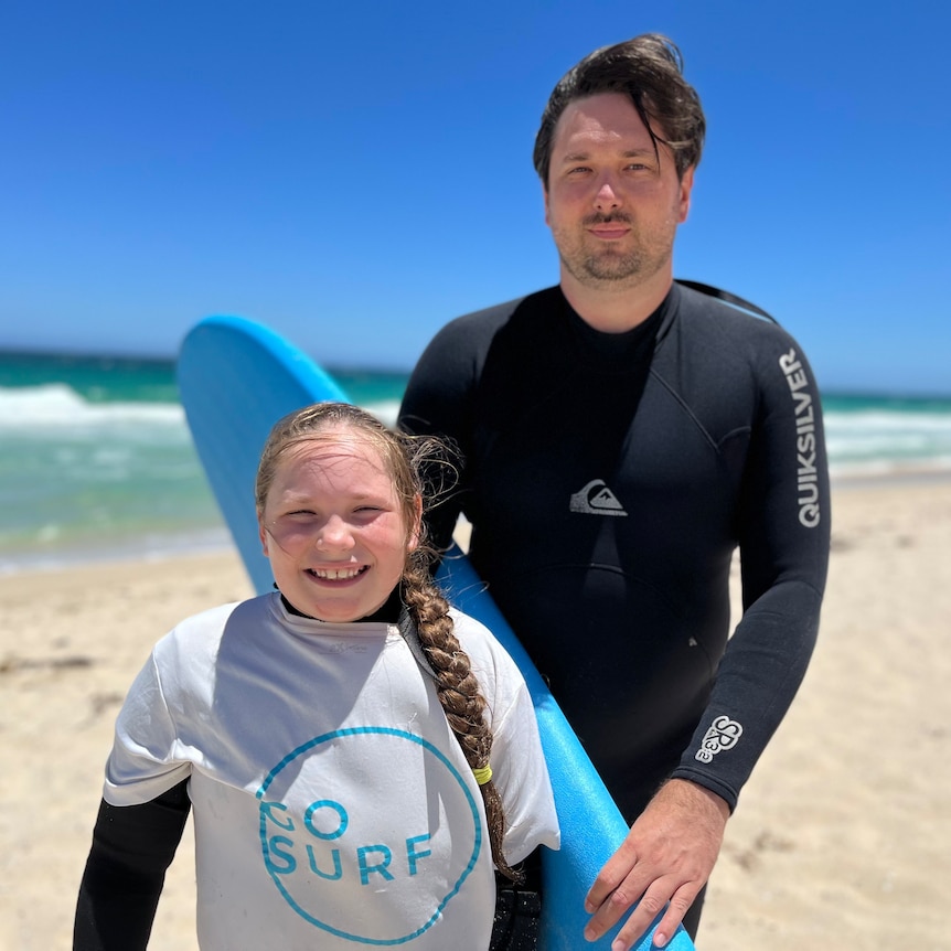 Young girl stands in front of her dad who is holding a surfboard at the beach