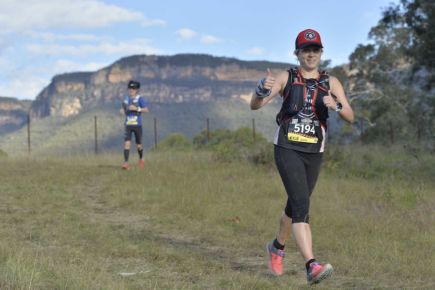 A woman gives the thumbs up as she runs in a field during an ultramarathon. Another woman can be seen in the background.