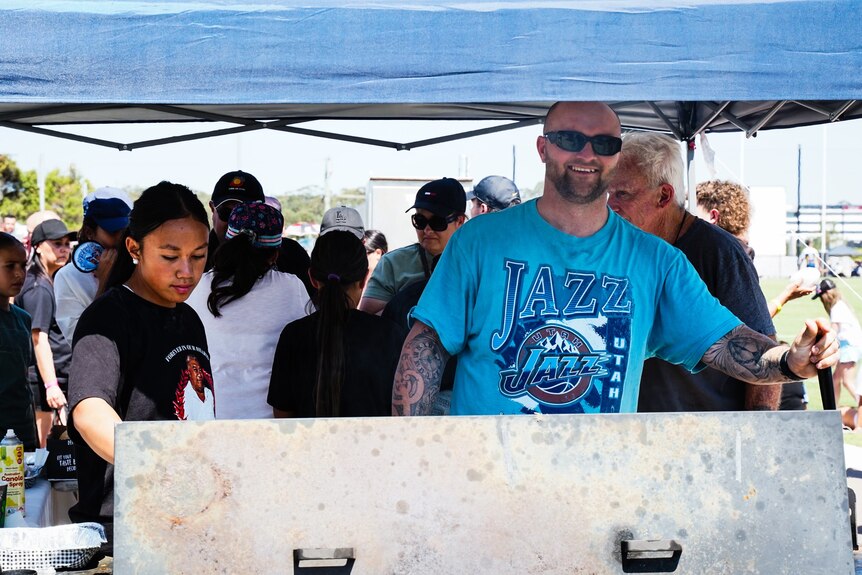 A young girl and a man standing behind a barbecue under a tent.