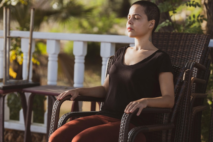 A woman with short brown hair meditates with her eyes closed while sitting on a chair