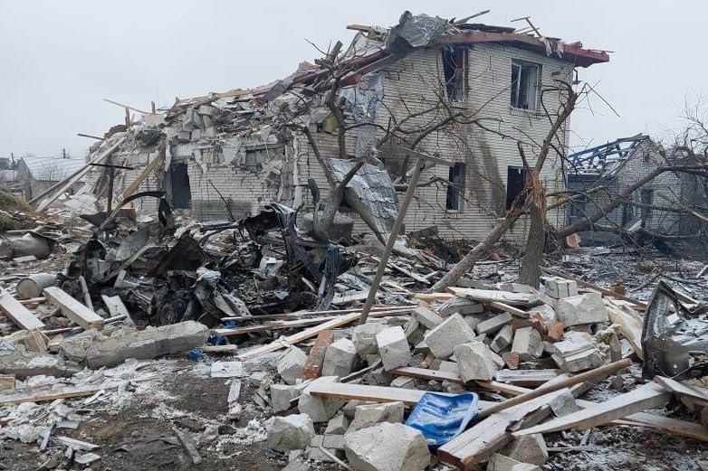 A crumpled stone building is surrounded by rubble and the burned-out remains of a car