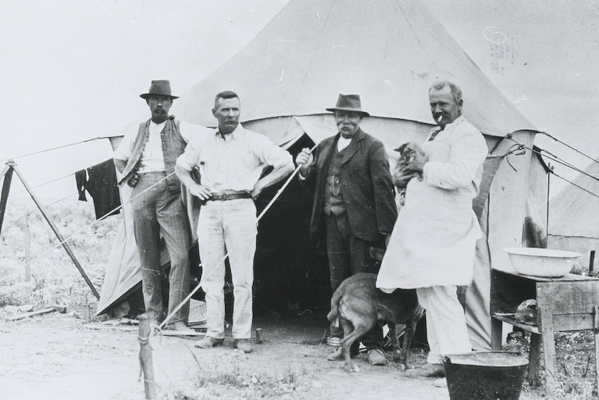 A black and white photo of a group of men standing in front of a tent.