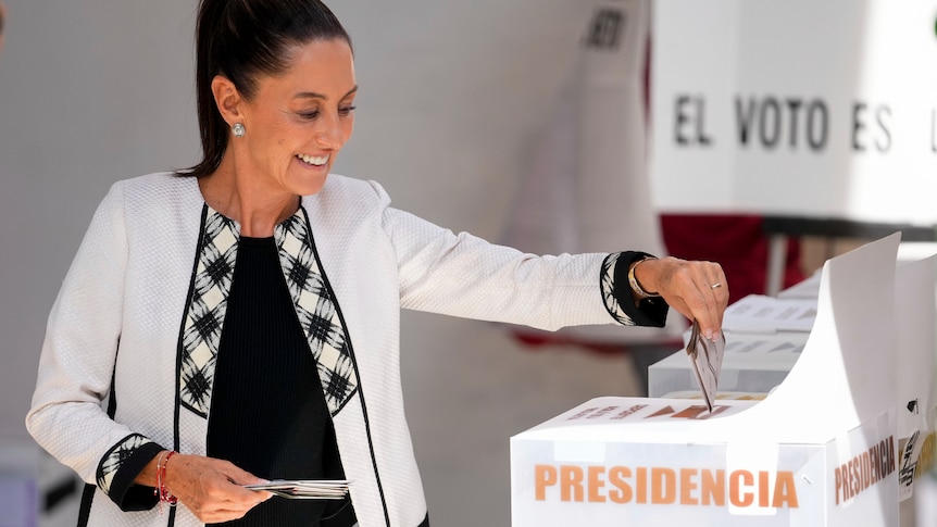 Claudia Sheinbaum, wearing a white tailored coat, places her vote in a box marked "Presidencia"