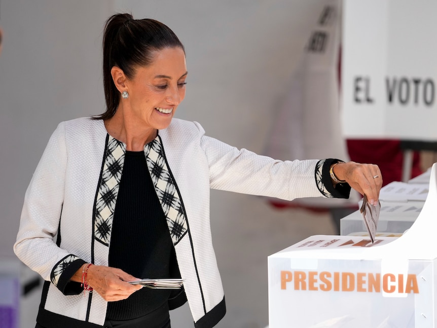Claudia Sheinbaum, wearing a white tailored coat, places her vote in a box marked "Presidencia"