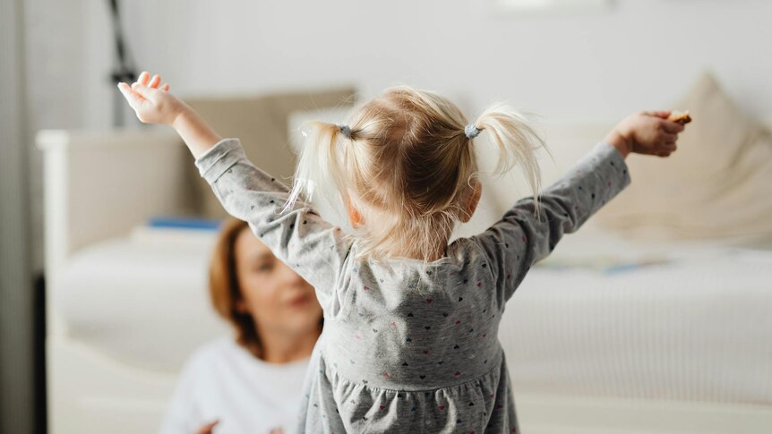 Photo of a small girl from behind - she is stretching her arms out in front of a woman who is watching