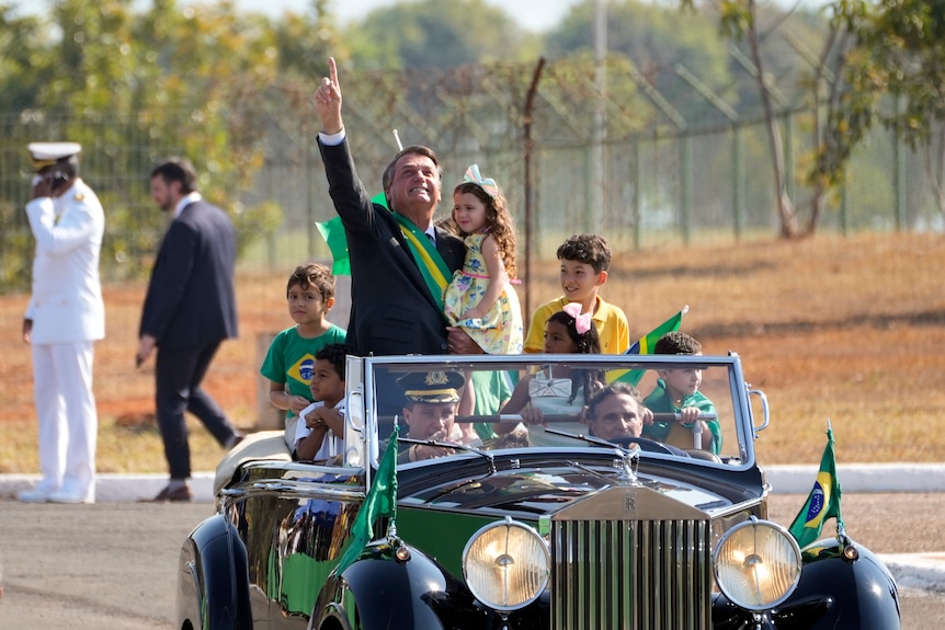 Jair Bolsonaro points to the sky as he rides in an open-top vintage car with a group of children