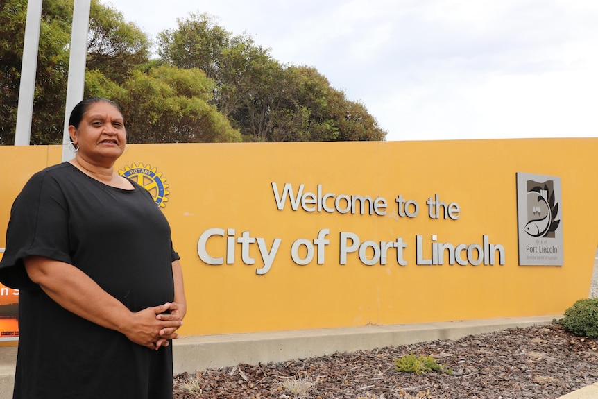 Emmalene Richards stands in front of a large yellow welcome to city of port lincoln sign on the highway. 