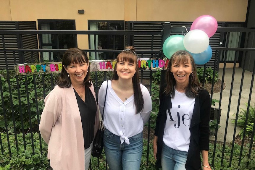 Three women stand in front of a fence with balloons and a 'happy birthday banner'.