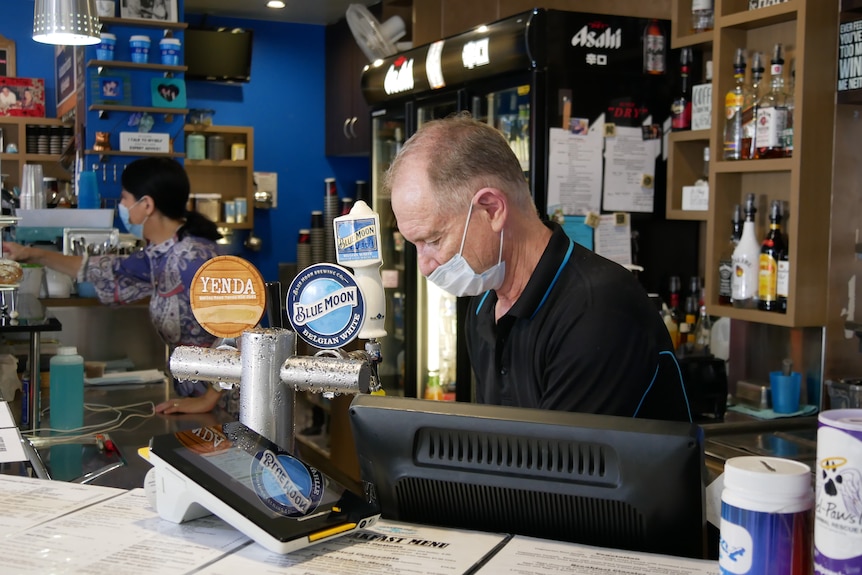 A man wearing a medical mask looks down behind the counter in a cafe.