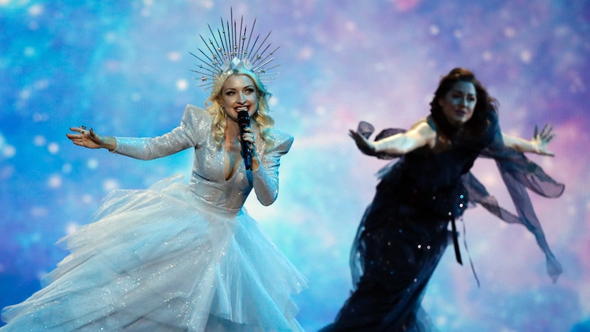 A singer in a ball gown with a crown on sings into a mike with a dancer reaching out behind her.
