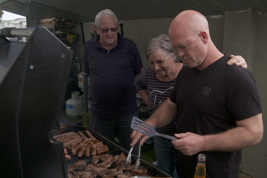 A bald man in black t-shirt stands cooking at barbecue next to his parents
