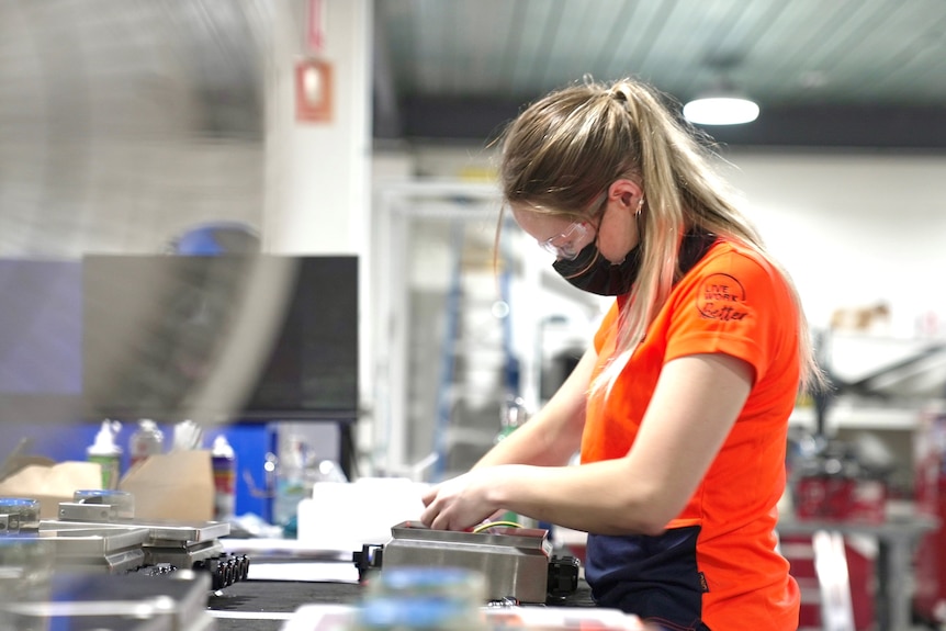 A young woman in a bright orange T-shirt works at a bench in a workshop. She has long, blonde hair.