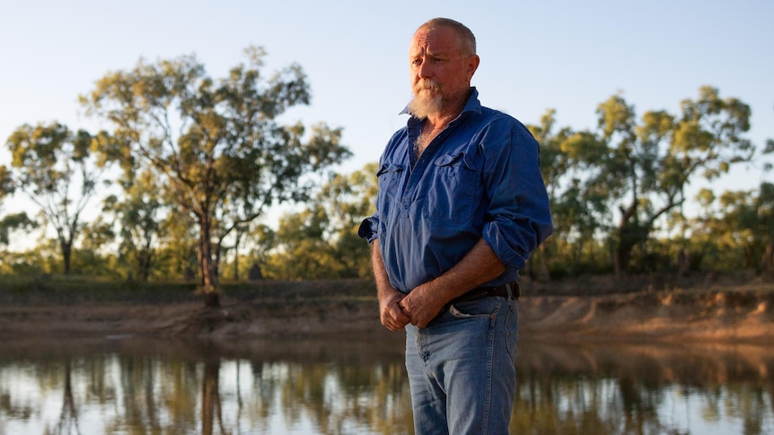 A middle-aged pastoralist wears jeans and a long-sleeved work shirt, and looks over a body of water at Gorrie Station. 