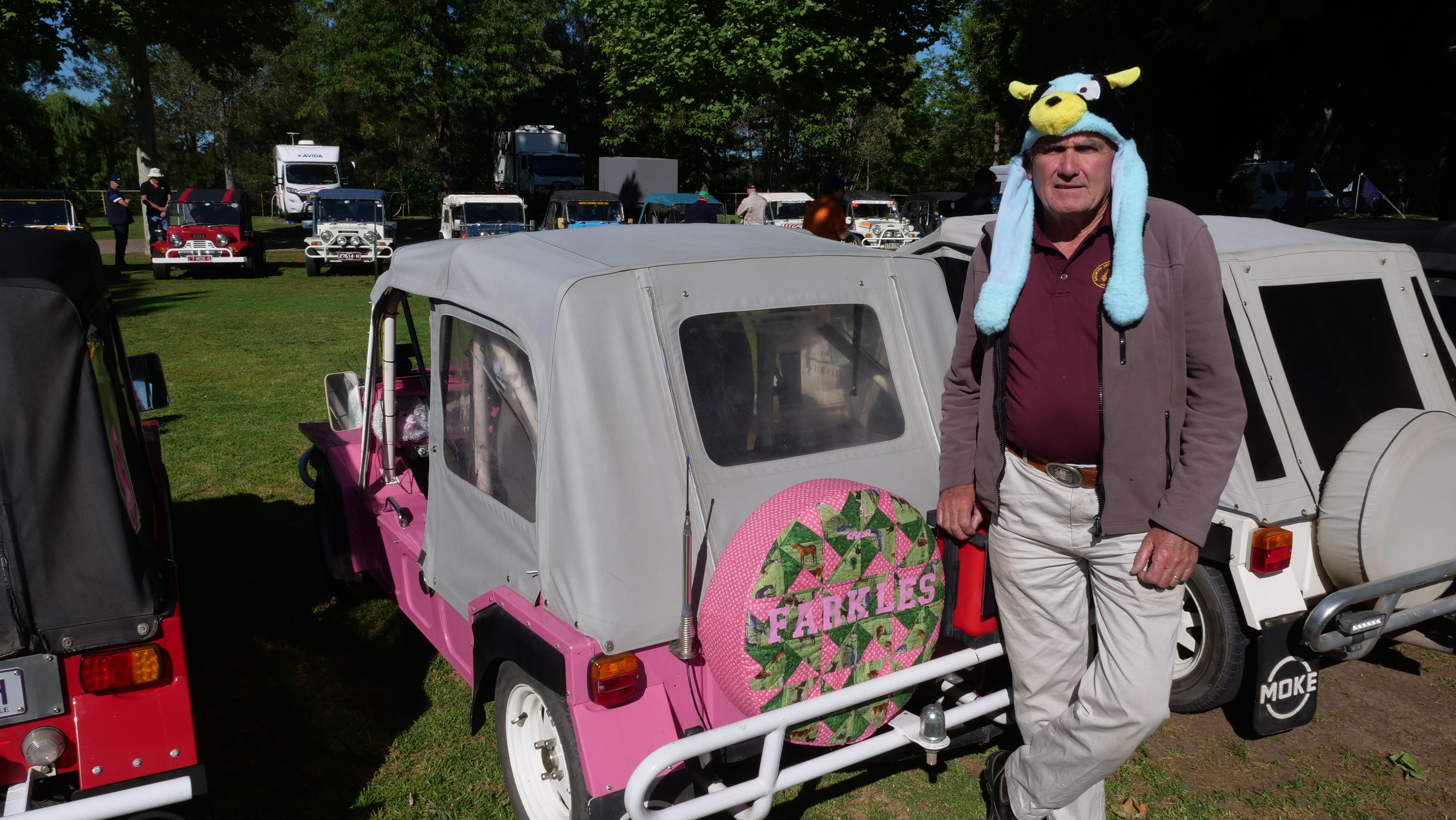 A man wearing a character hat stands in front of Mini Mokes including a pink one with Farkles written on the spare wheel cover.