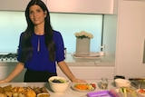 A woman stands in a kitchen with a spread of food in front of her.