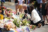 A suited man and a woman in a white blazer kneel to read messages on a floral tribute