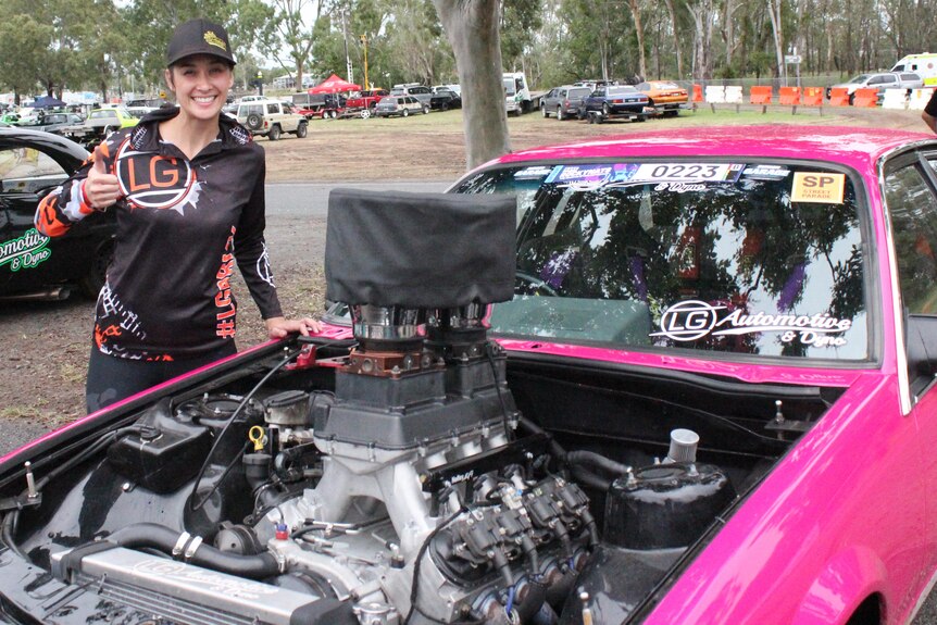 A woman stands next to her pink car with a revealed motor. She is smiling.