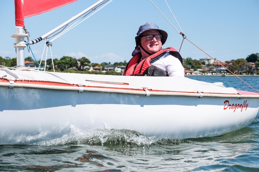 A man in a brimmed bucket hat and glasses wears a red lifejacket aboard a small sailboat